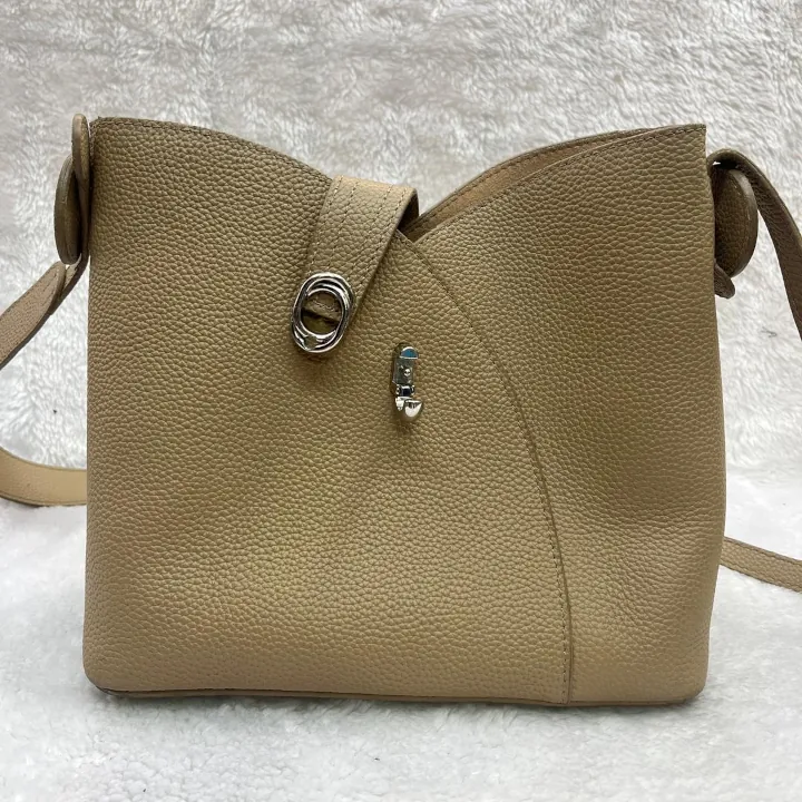 Beige pebbled leather crossbody bag with a curved top edge and a unique metal twist-lock closure.