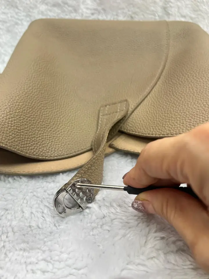 The worn twist closure on the strap is being removed from the beige leather hand bag by a screwdriver.