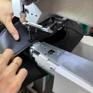 Close-up of hands using an industrial sewing machine to repair a leather bag, stitching along a seam to restore durability and appearance.