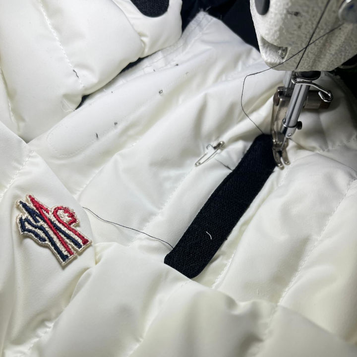 A close-up of a white Moncler winter jacket undergoing alteration with a sewing machine. The Moncler logo is visible on the fabric, while a black Velcro strap is being stitched. The sewing needle and thread are actively engaged in the tailoring process, ensuring a precise and seamless fit